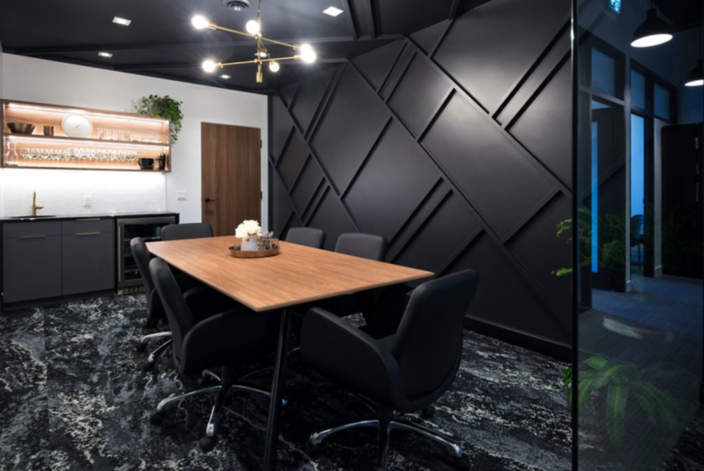 Modern office boardroom with feature wall in black with tone on tone geometric pattern, one white wall with light wood shelving and a light wood table with black chairs.